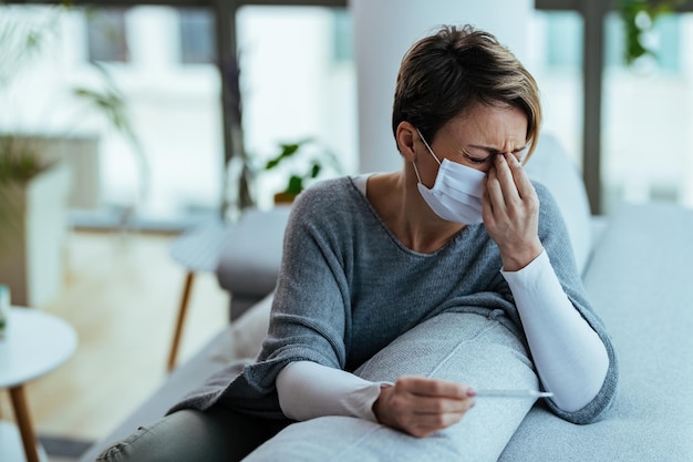 Foto giovane donna con maschera facciale che si sente male e tiene la testa dolorante mentre misura la temperatura a casa