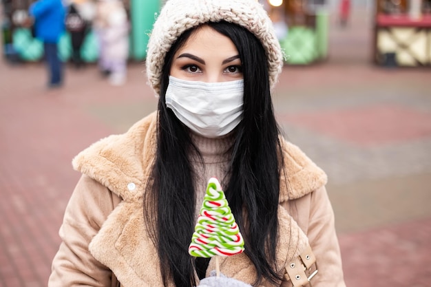Молодая женщина с маской для лица держит конфету, стоя на зимней праздничной ярмарке