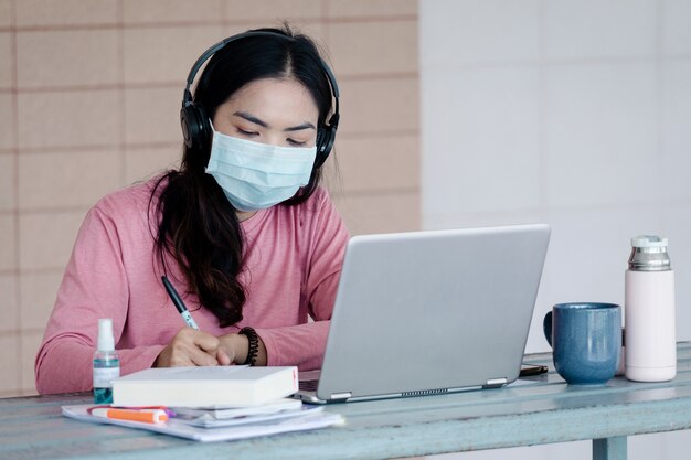 A young woman with face mask working on laptop at home during Coronavirus or COVID-19  pandemic.. An ault learner studying online at home. Stock photo