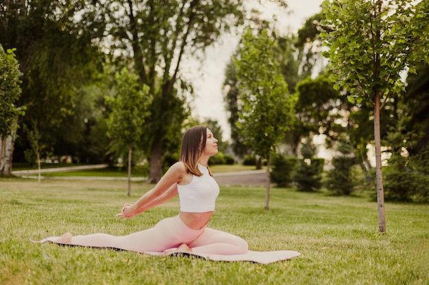 フィットフィギュアを持つ若い女性は、芝生の上の緑豊かな公園の体操マットでヨガを練習し、スポーツウェアの自然の背景でストレッチ体操を行います 健康的なライフスタイルと体との調和の概念