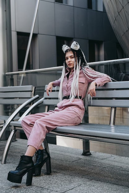 Young woman with futuristic looks sitting on a bench Girl with black and white pigtails Against the background of a futuristic building