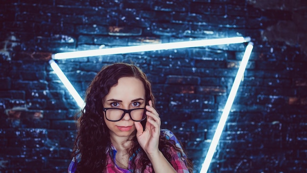 Young woman with glasses in colored plaid shirt posing on background shabby brick wall with glowing lamps Portrait of charming female holding eyeglasses with her hand against illuminated wall