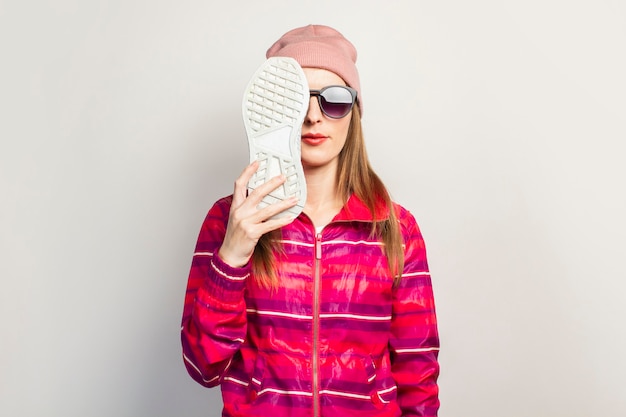 Young woman with glasses, hat and pink sports jacket with serious face covers her face with one sneaker