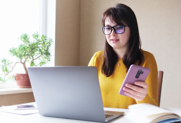 Young woman with glasses uses laptop and phone at home