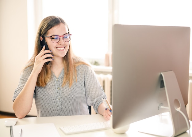 スマートフォンとコンピューターを使用して自宅で仕事をしている眼鏡の若い女性