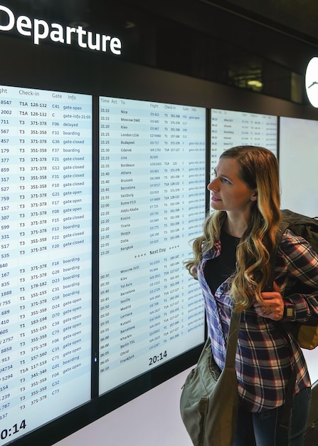 Young woman with green backpack and shirt looking at departure board screen at the airport