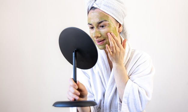 Young woman with a green mask on her face in a bathrobe in front of a mirror