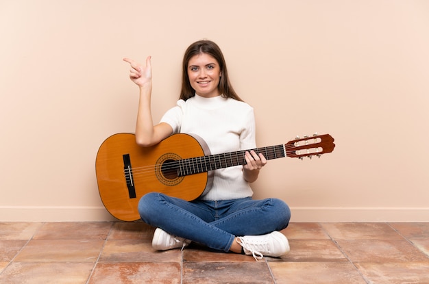 Young woman with guitar sitting on the floor pointing finger to the side