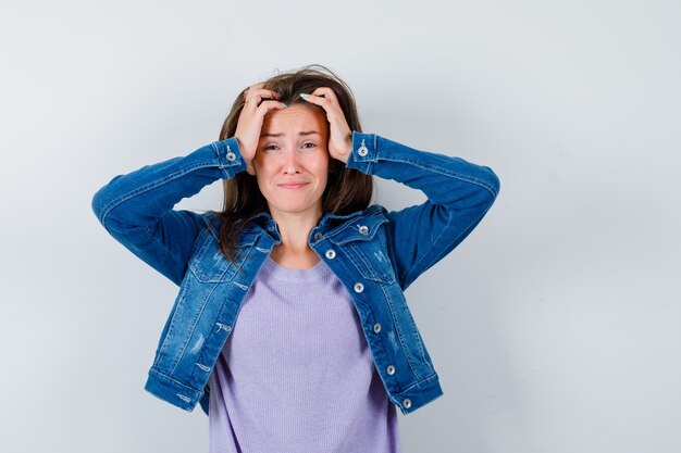 Young woman with hands on head in t-shirt, jacket and looking mournful , front view.