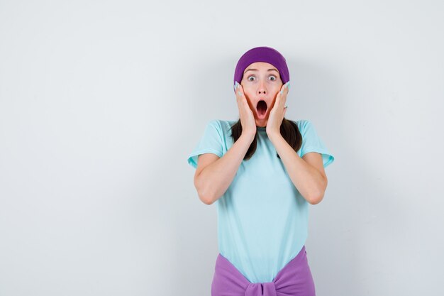 Молодая женщина с руками возле рта, широко открытым ртом в синей футболке, фиолетовой шапочке и удивленным видом. передний план.