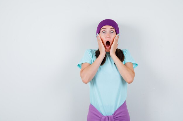 Young woman with hands near mouth, keeping mouth wide open in blue t-shirt, purple beanie and looking surprised. front view.