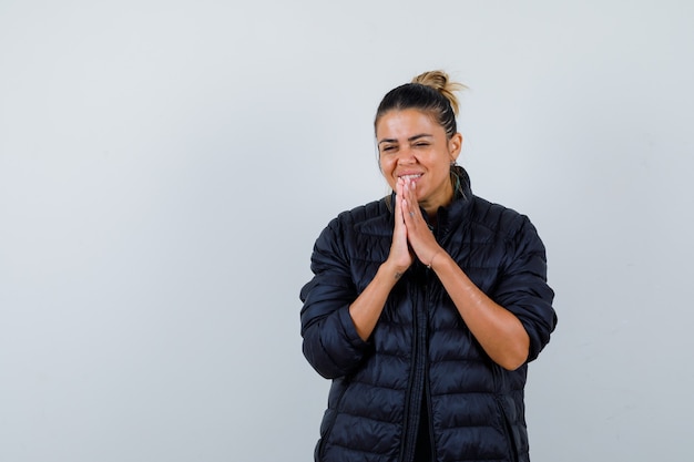 Young woman with hands in praying gesture in puffer jacket and looking cheery , front view.