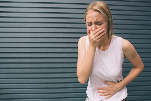 Young woman with hands on stomach having bad aches pain isolated on gray . Food poisoning, influenza, cramps. Health issues problems .