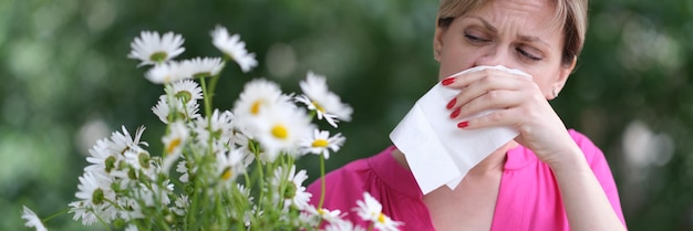 Young woman with hay fever blowing her nose in napkin near chamomile flowers