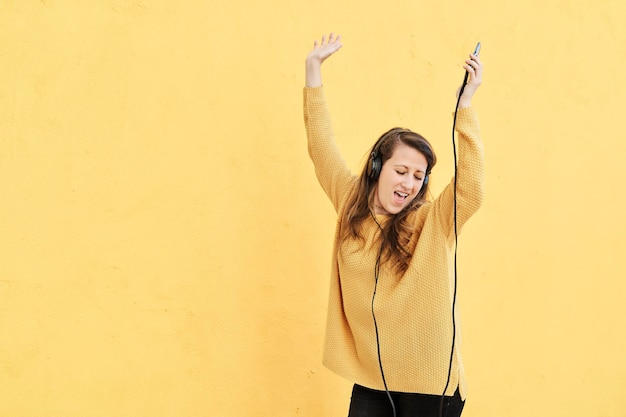 Young woman with headphones listening to music and dancing with her arms raised on a yellow background