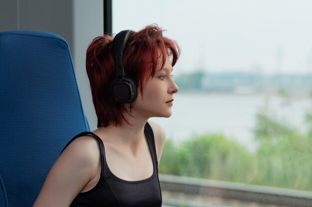 Young woman with headphones rides in a moving suburban train and looks out the window