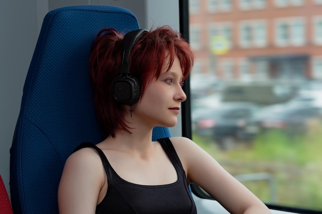 Young woman with headphones rides in a moving suburban train and looks out the window