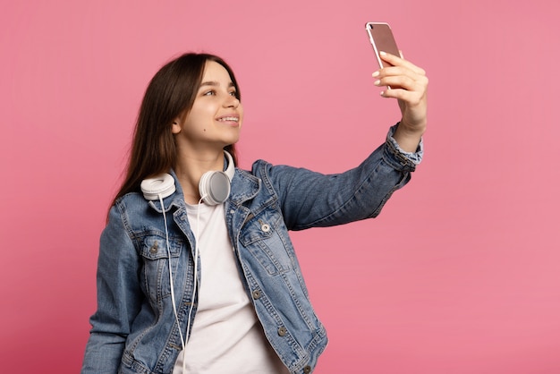 young woman with headphones, takes selfie, poses at camera isolated on pink