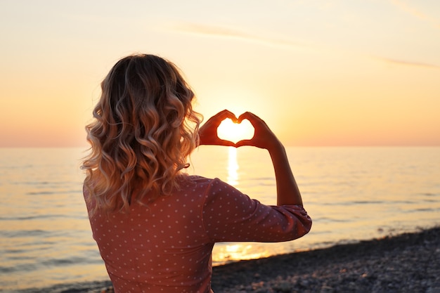 Young woman with heart-shaped fingers stands with her back on the seashore at sunset