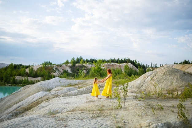 Молодая женщина с дочерью в желтом платье возле озера с лазурной водой и зелеными деревьями. Концепция счастливых семейных отношений