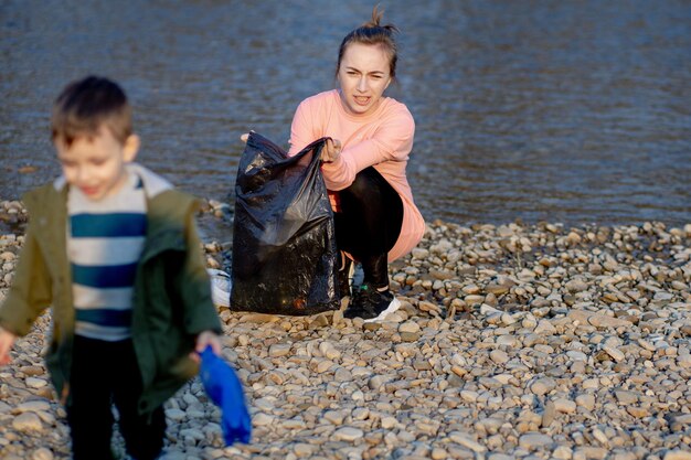 Photo young woman with her son collects plastic garbage in a garbage bag on the river empty used dirty plastic bottles environmental pollution shore