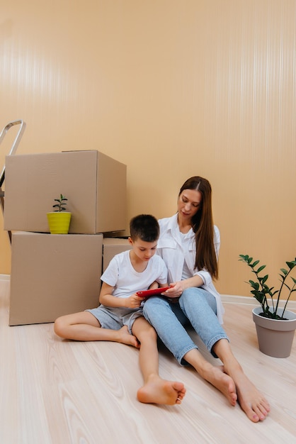 A young woman with her son is sitting in front of the boxes and rejoicing at the housewarming after moving in. Housewarming, delivery and freight transportation, purchase of real estate.