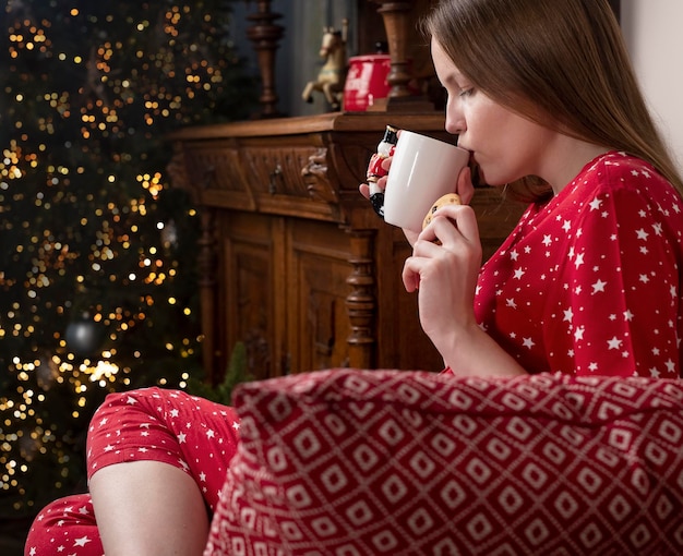 Young woman with hot tea cup sitting in chair at cozy home on\
winter holidays person in red pajamas ...