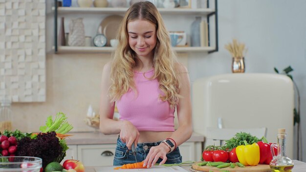 家庭のキッチンで木製のまな板に生のニンジンを刻むナイフを持つ若い女性健康的な食事と料理のコンセプト