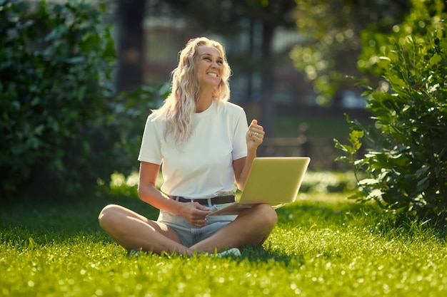 Young woman with laptop sits on the grass in the park on a sunny day.