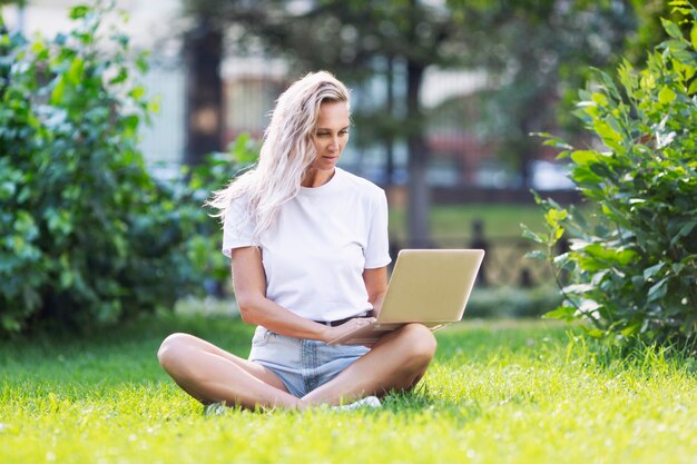 Young woman with laptop sits on the grass in a summer park.