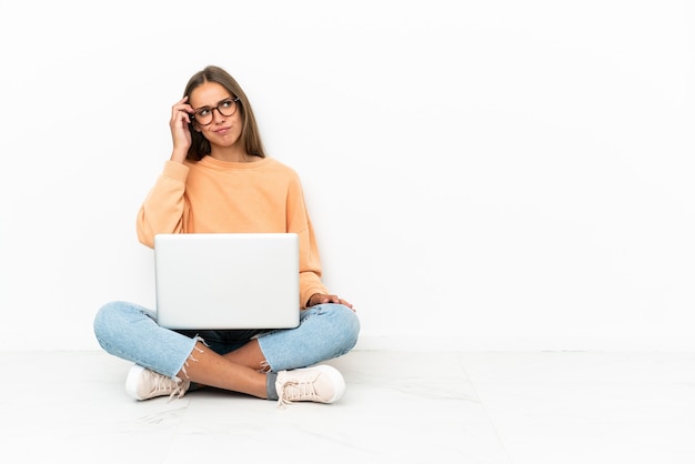 Young woman with a laptop sitting on the floor having doubts and with confuse face expression