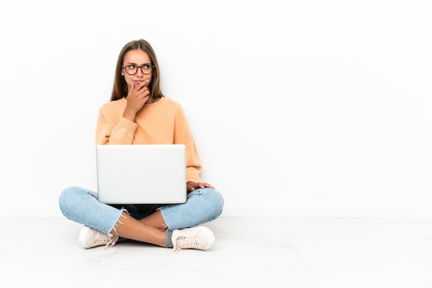 Young woman with a laptop sitting on the floor having doubts and with confuse face expression