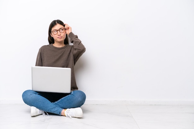 Young woman with a laptop sitting on the floor having doubts and with confuse face expression