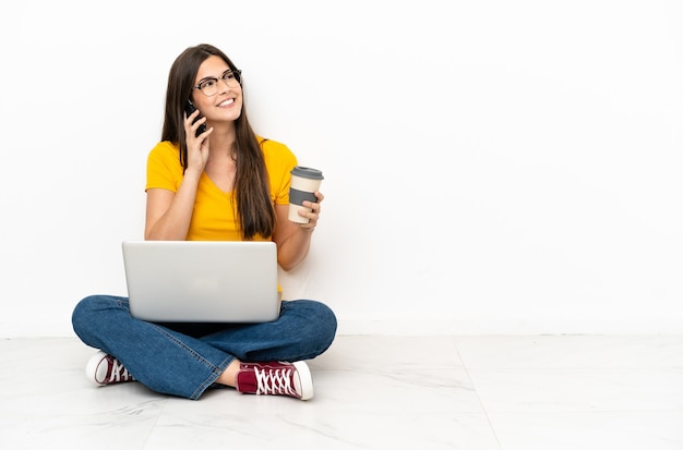 Young woman with a laptop sitting on the floor holding coffee to take away and a mobile