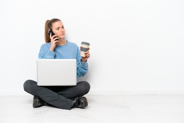 Молодая женщина с ноутбуком сидит на полу, держа кофе на вынос и мобильный