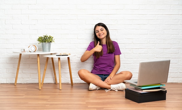 Молодая женщина с ноутбуком сидит на полу в помещении, показывая большой палец вверх