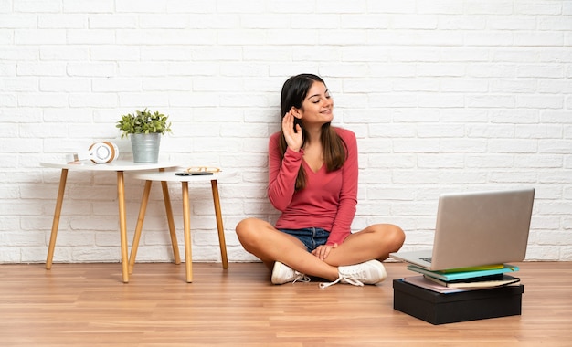 Young woman with a laptop sitting on the floor at indoors listening to something by putting hand on the ear