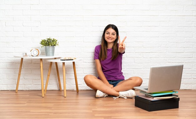 笑顔で勝利のサインを示して屋内で床に座っているラップトップを持つ若い女性