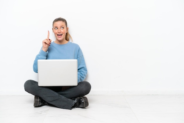 Young woman with a laptop sitting on the floor intending to realizes the solution while lifting a finger up