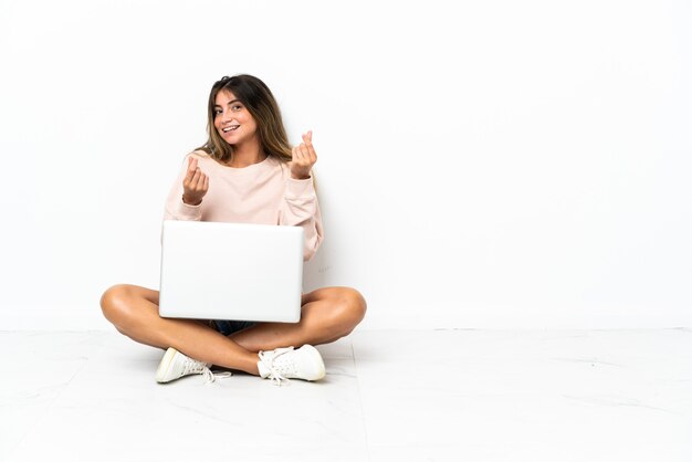 Giovane donna con un computer portatile che si siede sul pavimento isolato su fondo bianco che fa il gesto dei soldi