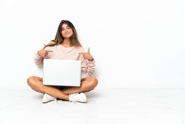 Young woman with a laptop sitting on the floor isolated on white background proud and self-satisfied