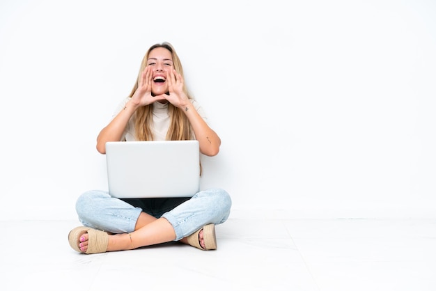 Young woman with laptop sitting on the floor isolated on white background shouting and announcing something