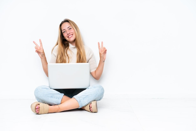 Photo young woman with laptop sitting on the floor isolated on white background showing victory sign with both hands