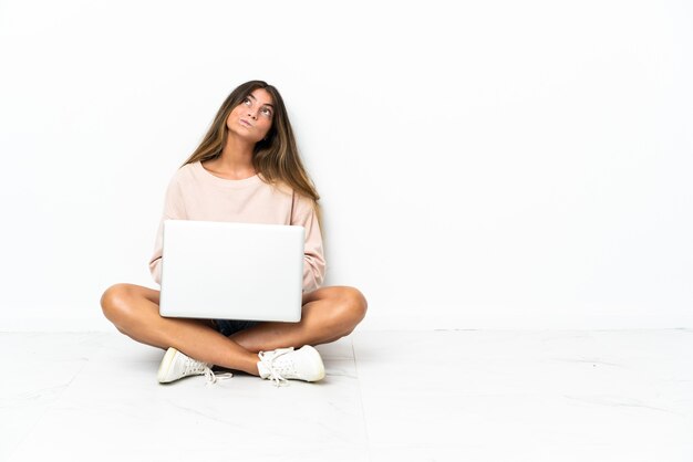 Молодая женщина с ноутбуком сидит на полу, изолированном на белой стене и смотрит вверх