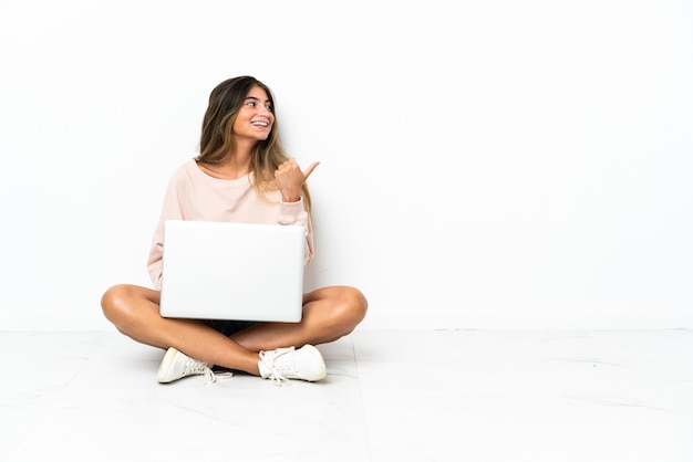 Young woman with a laptop sitting on the floor isolated on white wall pointing to the side to present a product