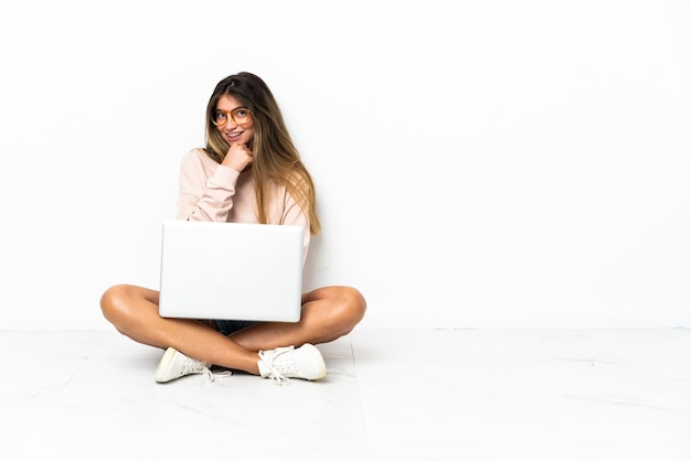 Young woman with a laptop sitting on the floor isolated on white wall with glasses and smiling