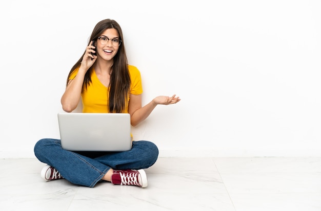 Young woman with a laptop sitting on the floor keeping a conversation with the mobile phone with someone