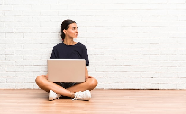 Giovane donna con un computer portatile che si siede sul pavimento che osserva al lato