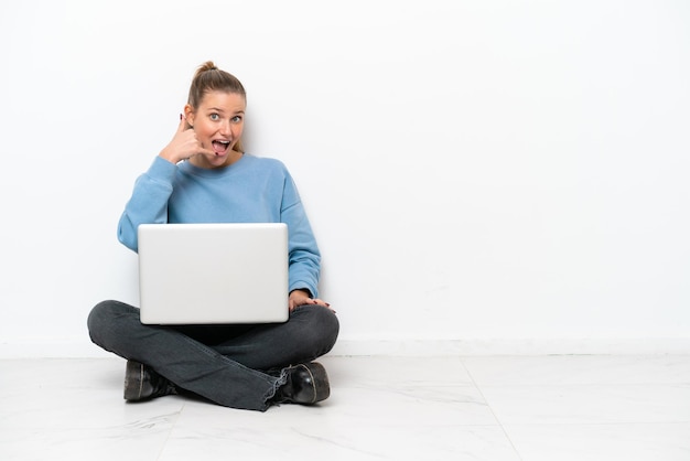 Young woman with a laptop sitting on the floor making phone gesture Call me back sign