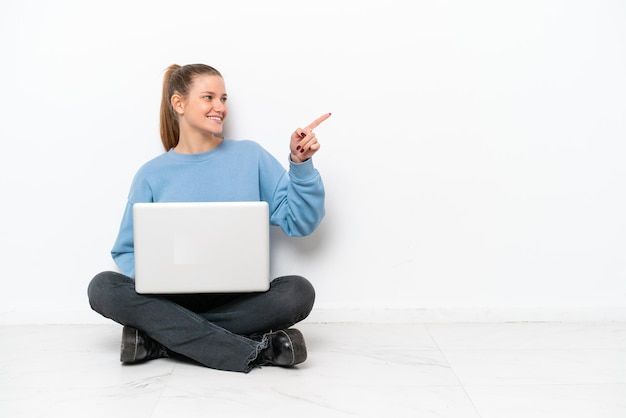 Young woman with a laptop sitting on the floor pointing finger to the side and presenting a product